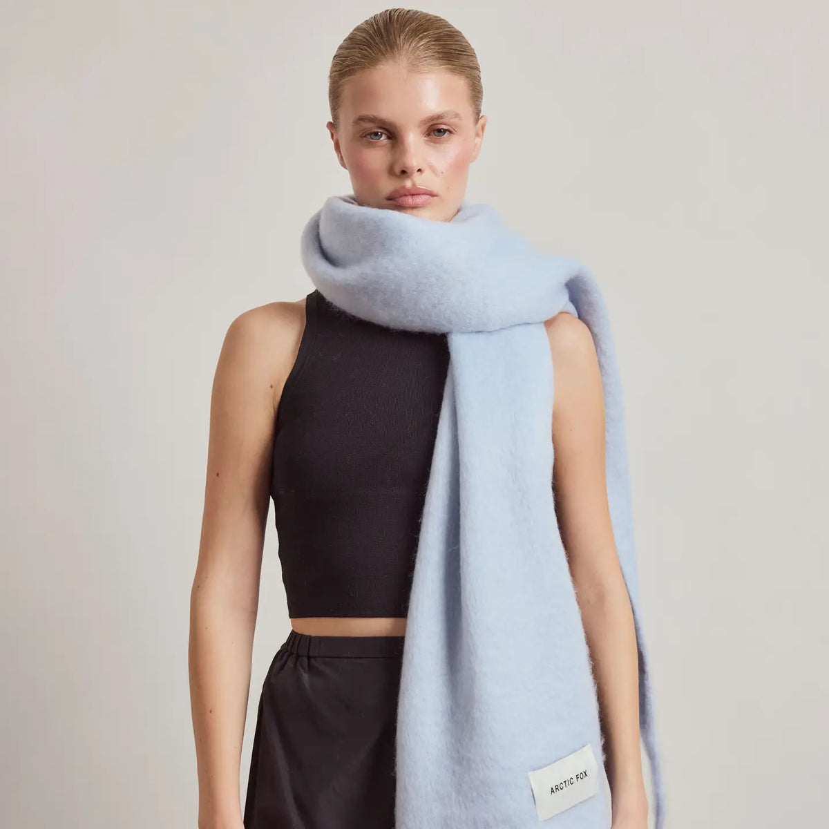 A model wearing a light blue fuzzy wrap scarf with a white brand label that reads 'Arctic Fox', styled over a black crop top and black pants against a neutral light gray background.