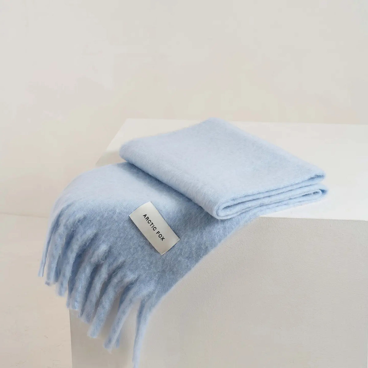 A folded light blue knit scarf with fringe, draped over a white display block. A small white label on the scarf reads 'Arctic Fox'.