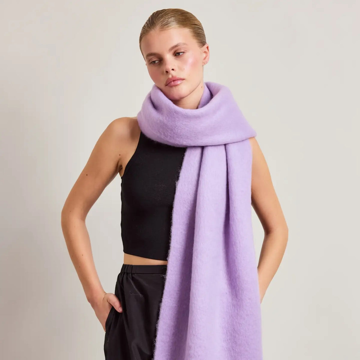 A model wearing a thick, soft lavender-purple fuzzy scarf wrapped around her neck, paired with a black sleeveless crop top and black pants, against a plain light gray studio background.