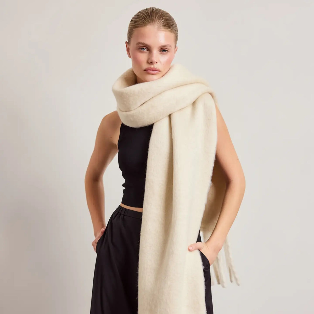 A woman modeling a thick, soft cream-colored knit scarf wrapped around her shoulders, paired with a black sleeveless top and black pants, against a plain light gray studio background.