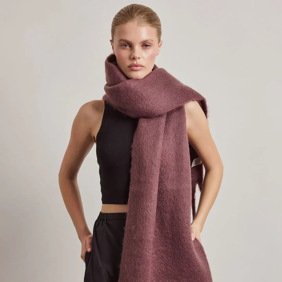 A model wearing a thick, fuzzy dusty rose scarf wrapped around her neck, paired with a black crop top and black pants, against a plain light gray studio background.