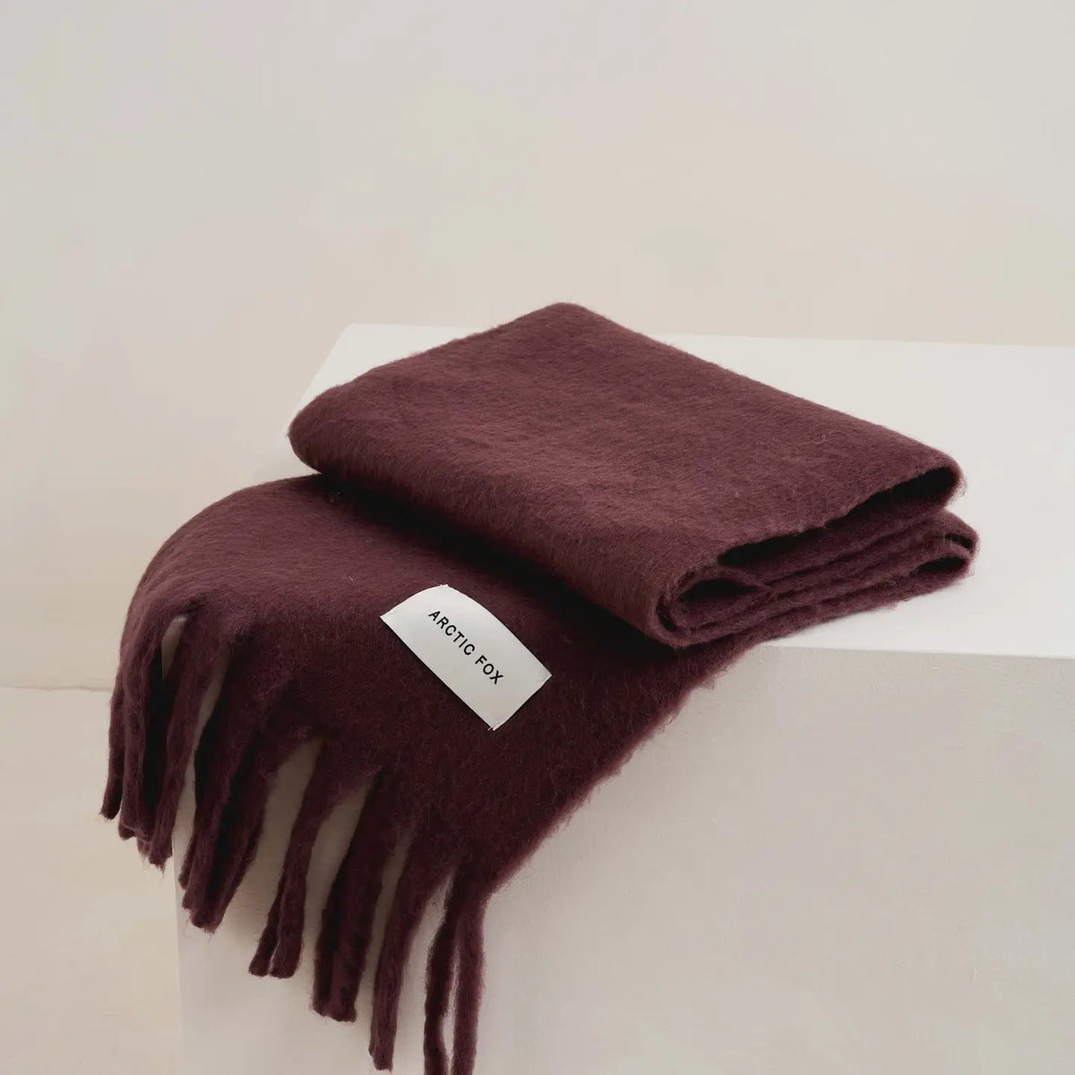 A folded dark plum-colored wool scarf with fringe, displayed on a white block against a light beige background. A white fabric label with the brand name 'ARCTIC FOX' is attached to the scarf.