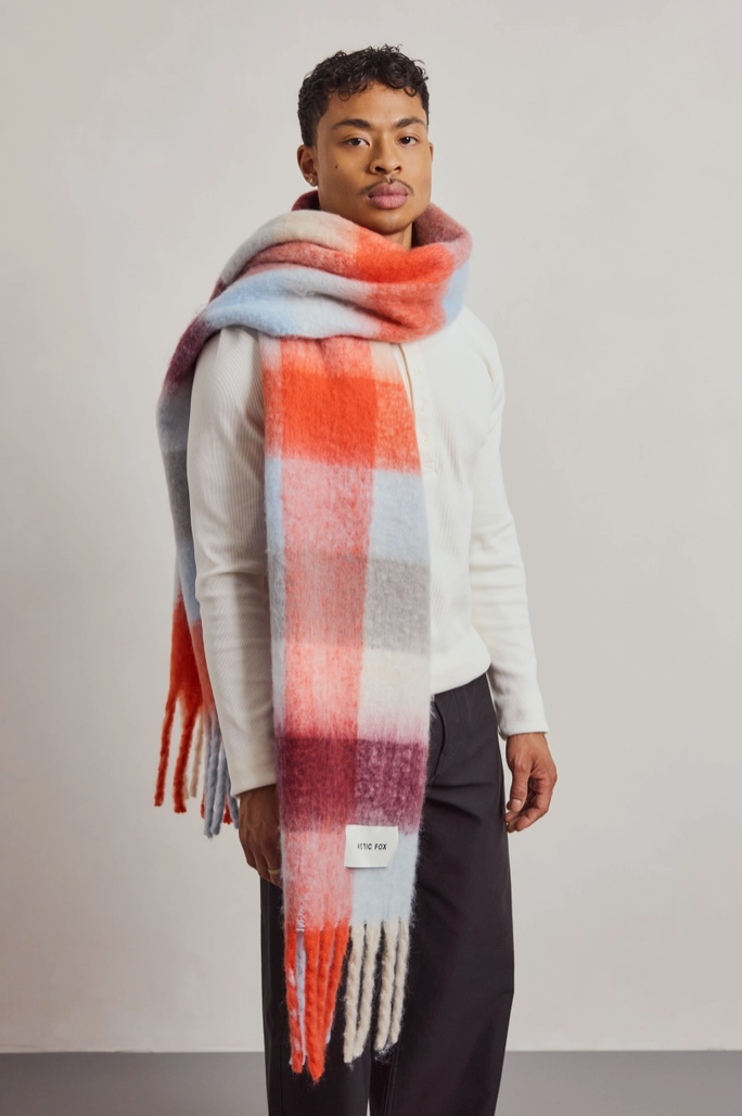 A man wearing a soft, oversized plaid scarf in orange, red, pink, blue, and white, paired with a white sweater and dark trousers, standing against a neutral light gray studio background.