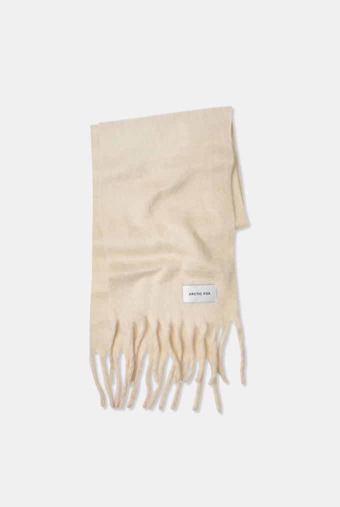 A light beige knit scarf with fringed ends and a small white rectangular brand label, photographed on a plain light gray background.